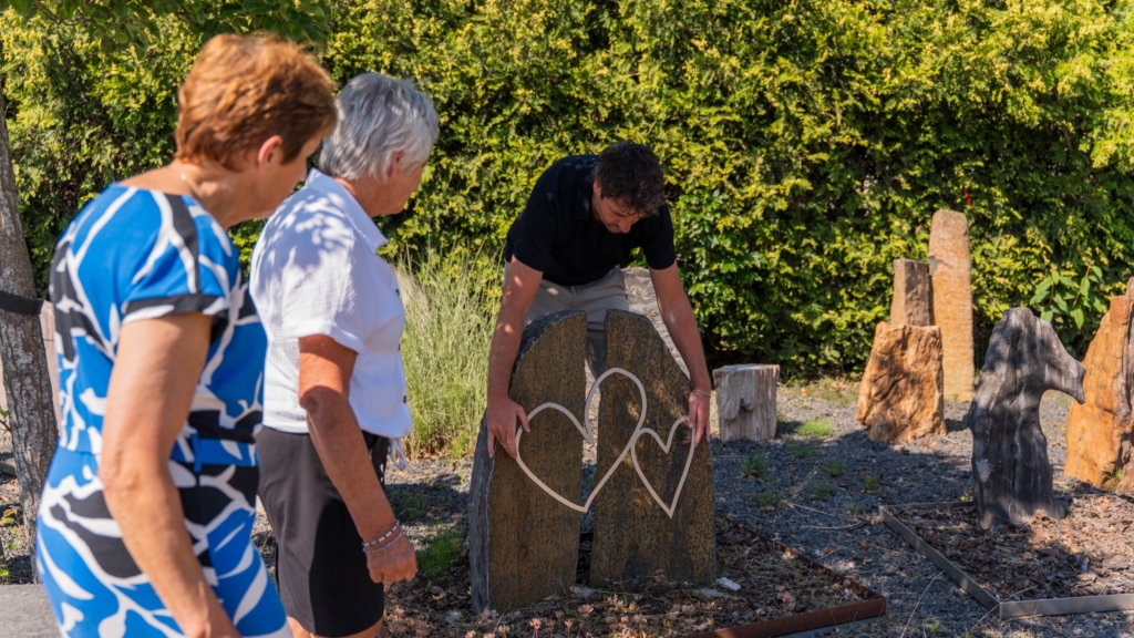 Adviseur van Hutting Natuursteen toont een hartornament aan twee klanten in de monumententuin